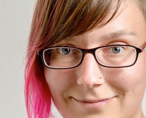 An image of Uta Hinrich, a middle aged woman with assymetrical brown and pink tipped hair, wearing glasses, with a nose piercing.