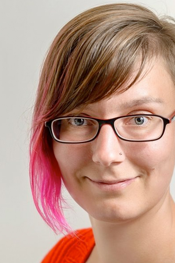 An image of Uta Hinrich, a middle aged woman with assymetrical brown and pink tipped hair, wearing glasses, with a nose piercing.