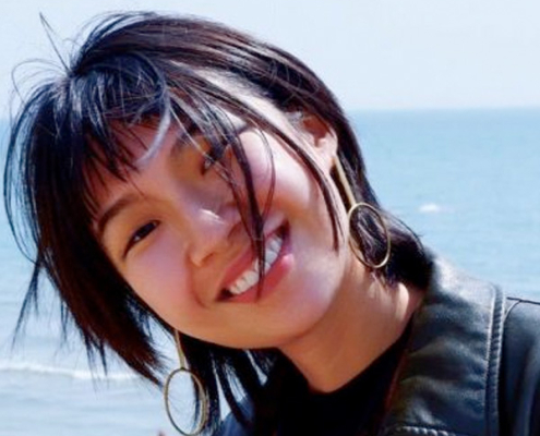 A picture of Yunung Chen, a young woman with should length black hair with a fringe, wand statement earrings, wearing a leather jacket standing on a pebble beach.